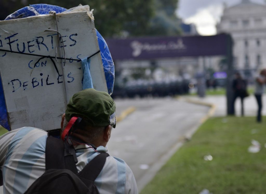 Foto de una mujer de espaldas sosteniendo un cartel en una manifestación. En el cartel se lee: "Fuertes con los débiles". Fuente: tapa del libro.